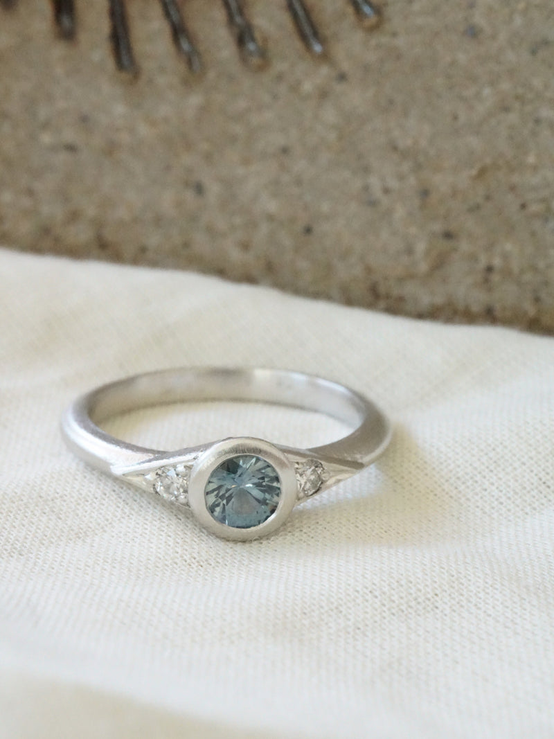 Shown: BIZARRE X -$1445 - Dahlia 3 Stone Ring - 0.25ct Aqua Montana Sapphire with 2mm diamond sides. Platinum. Smooth texture. Matte finish. Size 4.25. 